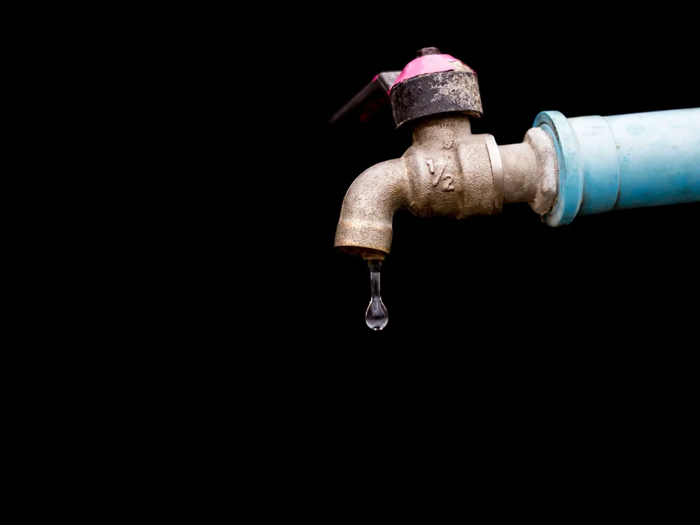 Water dripping from a worn brass tap against a black background